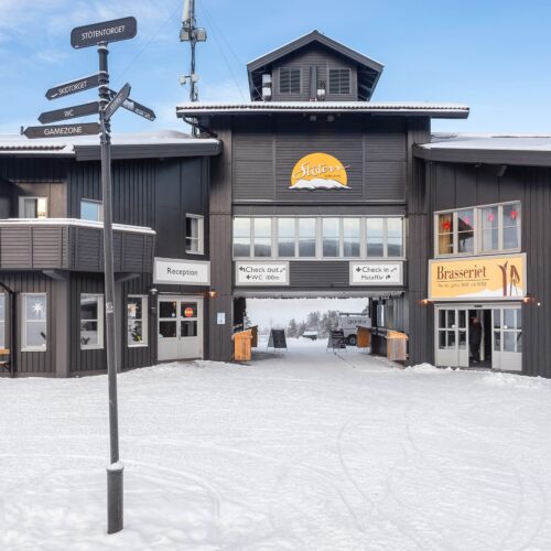Sort bygning med skilte til reception, skipas og brasserie, snedækket jord, vejskilt med anvisninger og åbning til skiområdet i baggrunden.