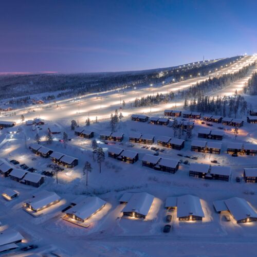 Snow-covered mountain village with illuminated ski slopes, wooden cabins and parked cars, surrounded by forest in the blue hour.