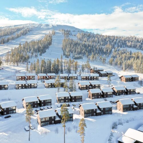 Snedækket bjerglandsby med rækker af brune træhuse, parkerede biler og omgivende grantræer, skiløjper i baggrunden under klar blå himmel.