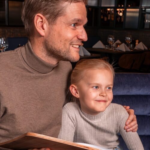 Person håller meny vid dukat bord i restaurang med glas, servetter och varm belysning.