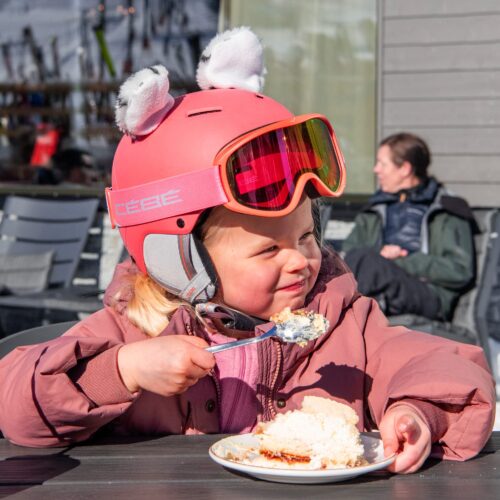 Person in pink ski jacket and helmet with ears eating waffle with cream on sunny outdoor terrace.