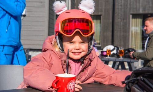 Person in pink ski jacket and helmet with ears drinking hot drink on sunny outdoor terrace.