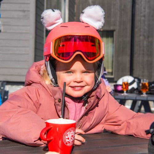 Person i rosa skidjacka och hjälm med öron dricker varm dryck på solig uteservering.