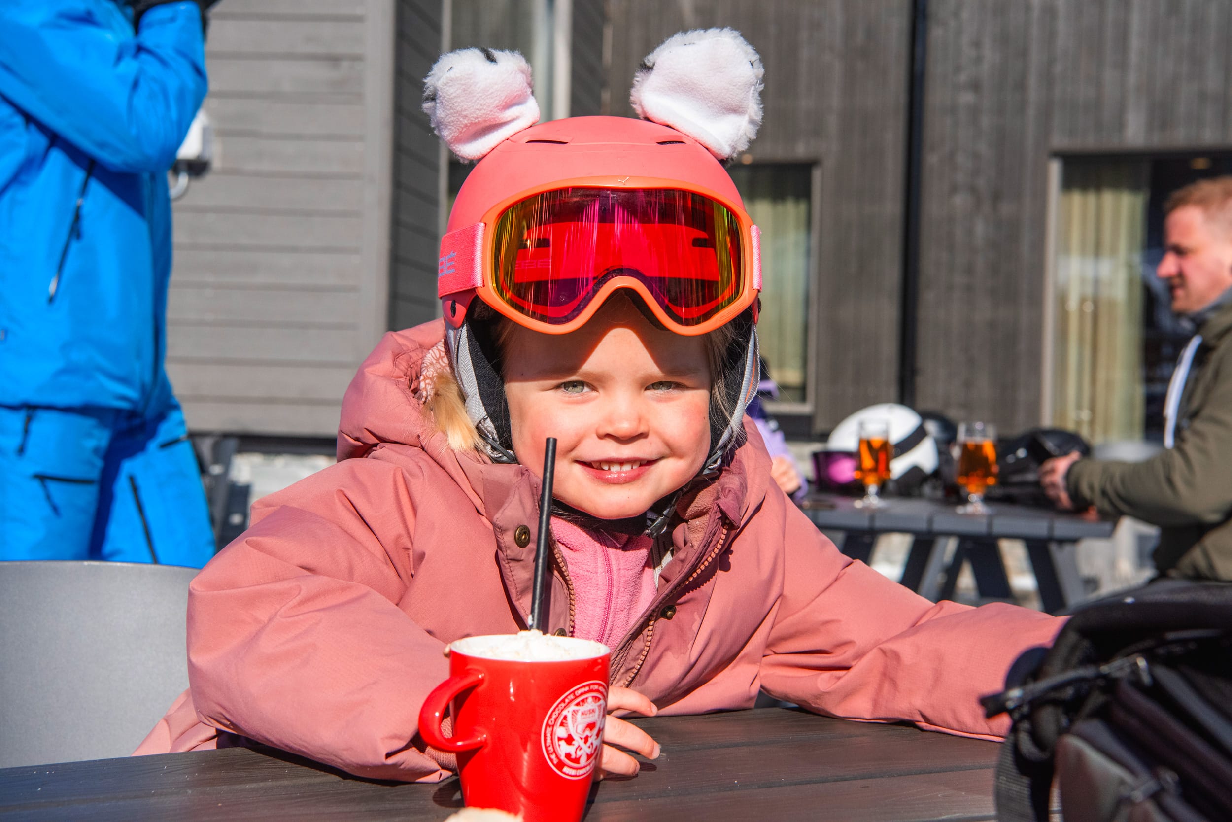 Person i rosa skidjacka och hjälm med öron dricker varm dryck på solig uteservering.