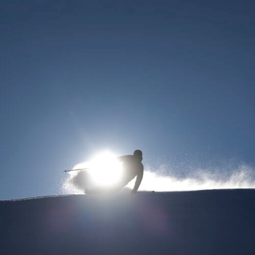 Silhouette of skiers swinging in powder snow on mountainside with bright sun behind