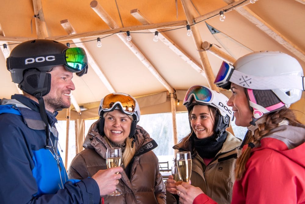 Fyra personer i skidutrustning skålar med glas mousserande vin inne i restaurangkåta med ljusslingor.