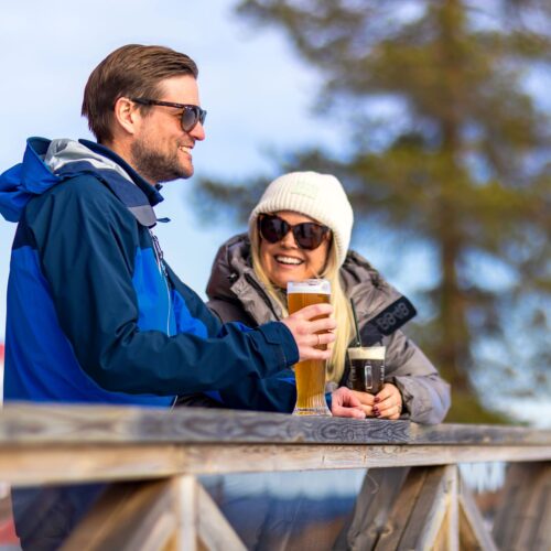 Two people stand by wooden fence on sunny terrace holding glasses of beer and dark drink.