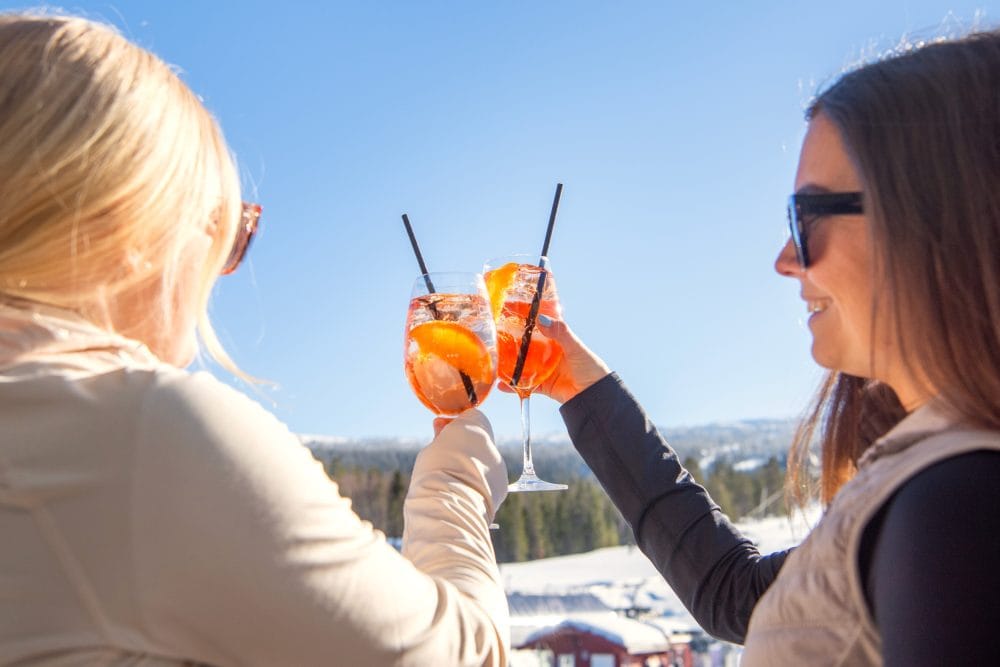 To mennesker skåler i orange drinks med sugerør mod en solrig skibakke og blå himmel.