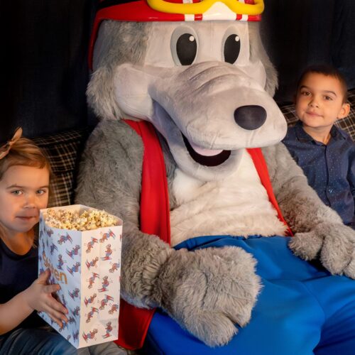 Vargy the mascot sitting on a sofa with children and a big box of popcorn