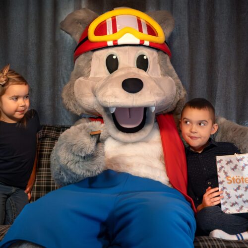 Vargy the mascot in red helmet and waistcoat sitting on sofa with kids and popcorn