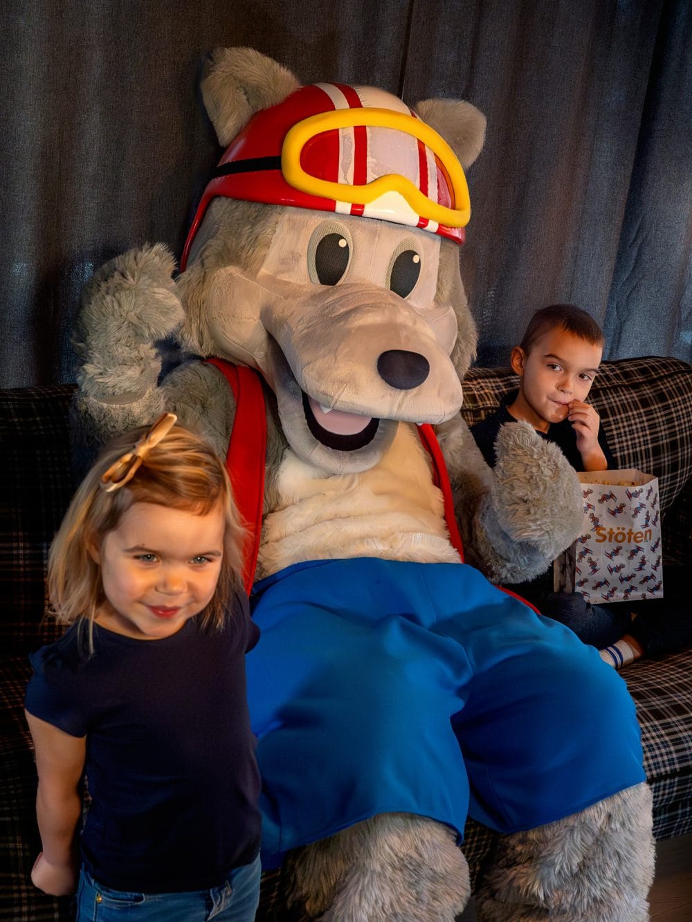Maskoten Vargy i röd hjälm och väst sitter i soffa med barn och popcorn