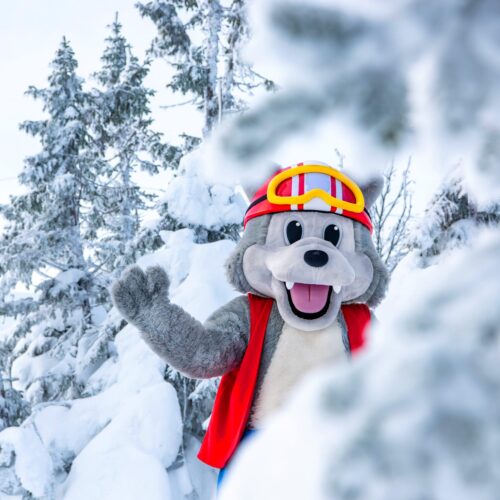 Maskotten Vargy i rød vest og skibriller står blandt snedækkede grantræer og vinker glad.