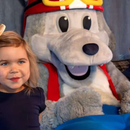 Maskoten Vargy i röd hjälm och väst sitter i soffa med barn och popcorn