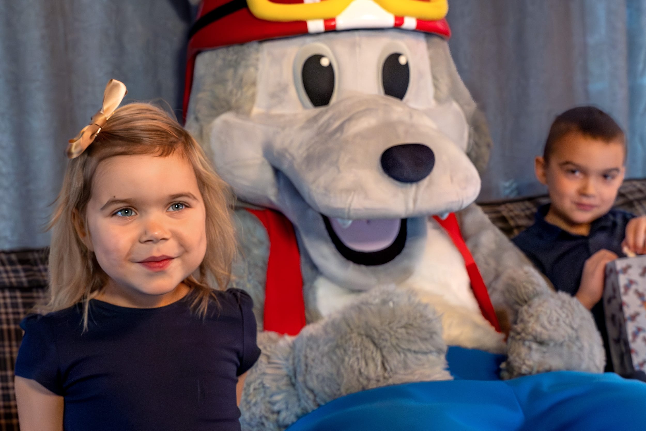 Maskoten Vargy i röd hjälm och väst sitter i soffa med barn och popcorn