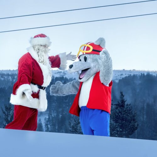 Maskoten Vargy i röd väst hälsar på jultomten i snöig skidbacke med lift i bakgrunden