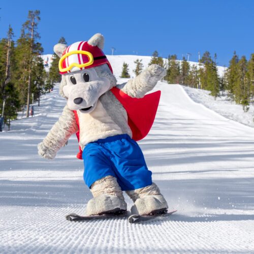 Maskotten Vargy i rød vest står på ski på en solrig piste med en skov i baggrunden.