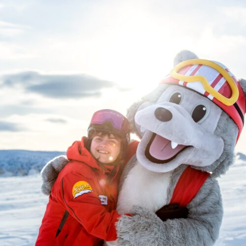 Vargy the mascot in a red waistcoat and ski goggles hugs a ski instructor in a snowy landscape.