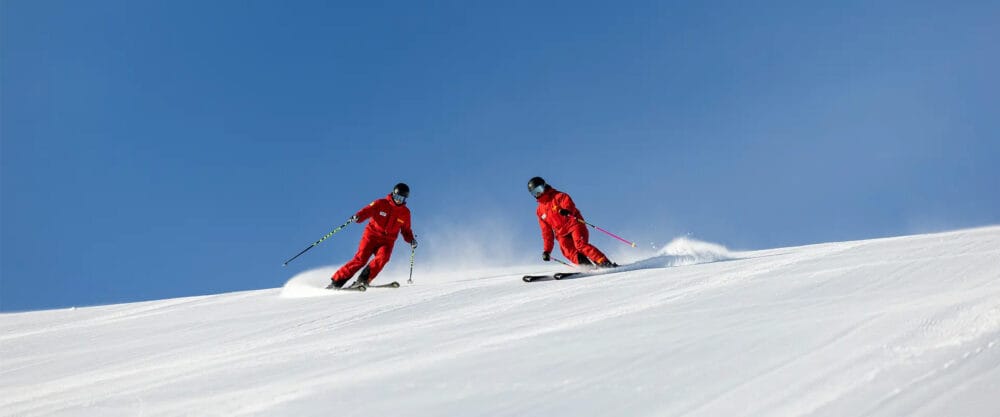 To skiløbere i rødt tøj svinger sig ned ad en snedækket piste under en klar blå himmel.