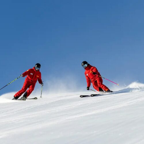Two skiers in red clothes swing down a snow-covered piste under a clear blue sky