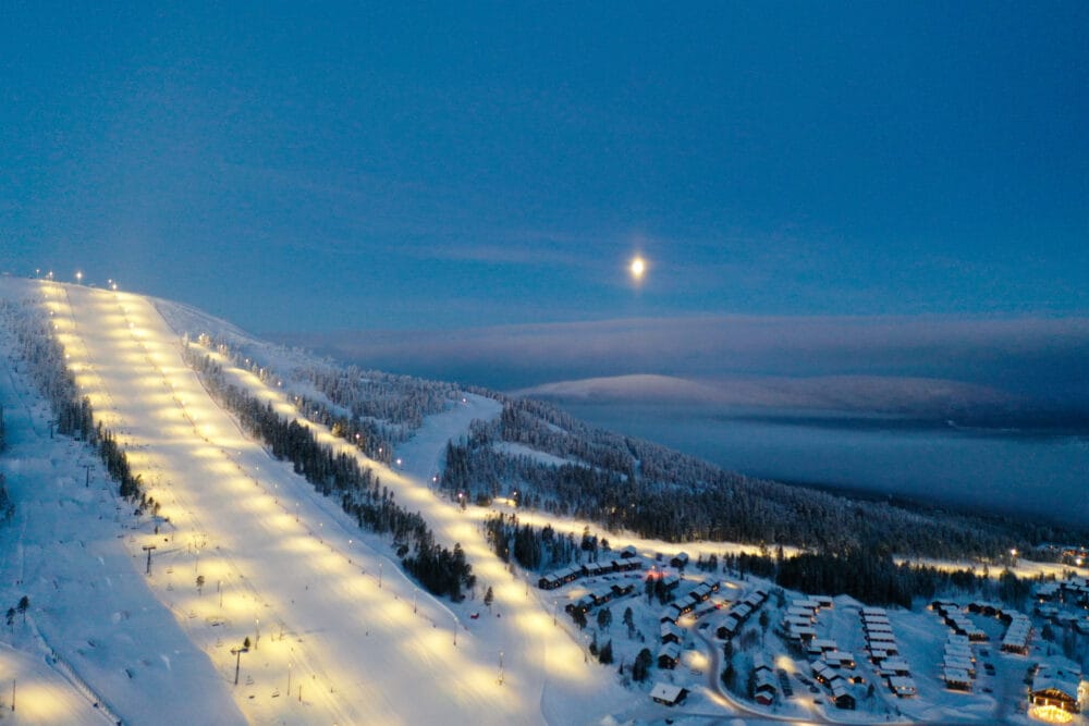 Upplyst skidbacke i kvällsmörker, snöklädda träd, måne över fjäll och liftområde.