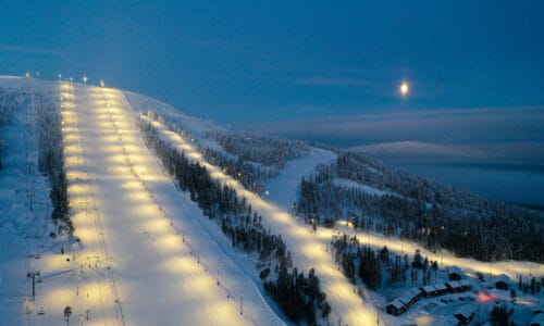 Illuminated ski slope in the evening darkness, snow-covered trees, moon over mountains and lift area.