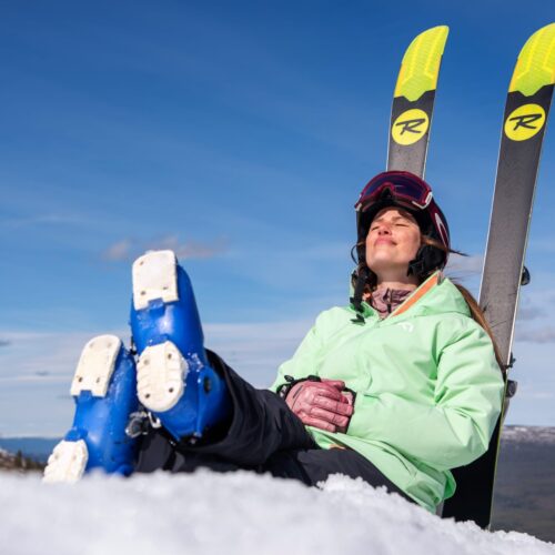 Person i lysegrøn jakke sidder i sneen med ski bag ryggen under blå himmel