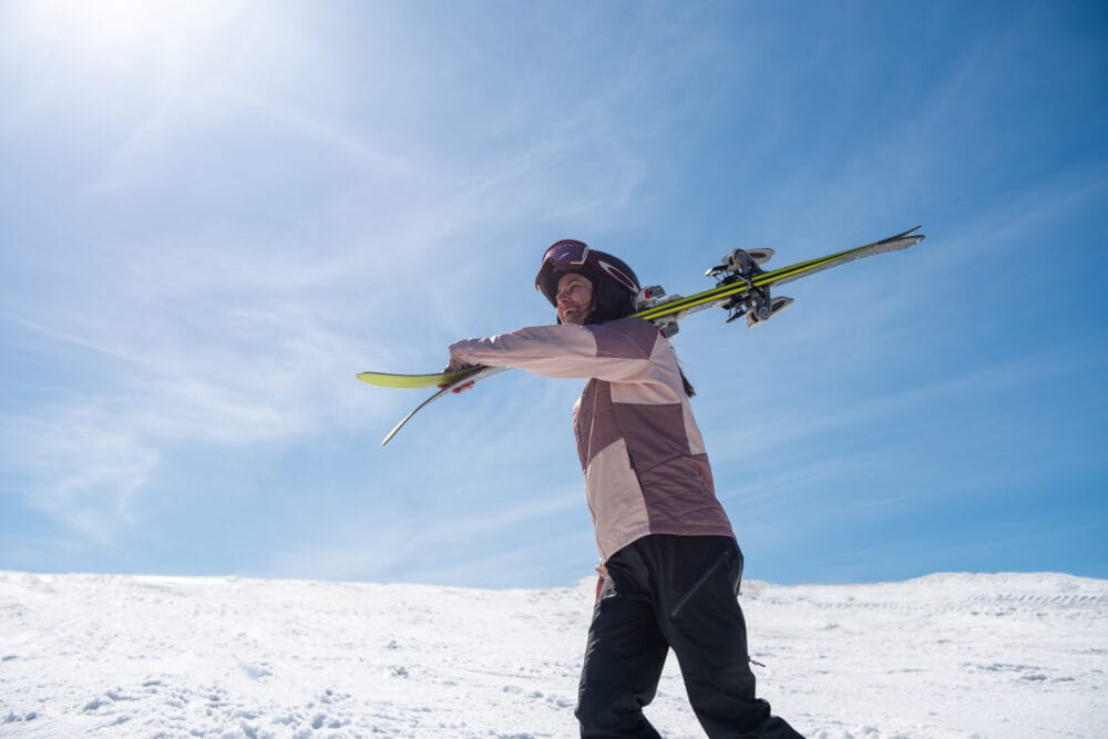 Person carrying skis over shoulder on snow-covered mountain under clear blue sky.