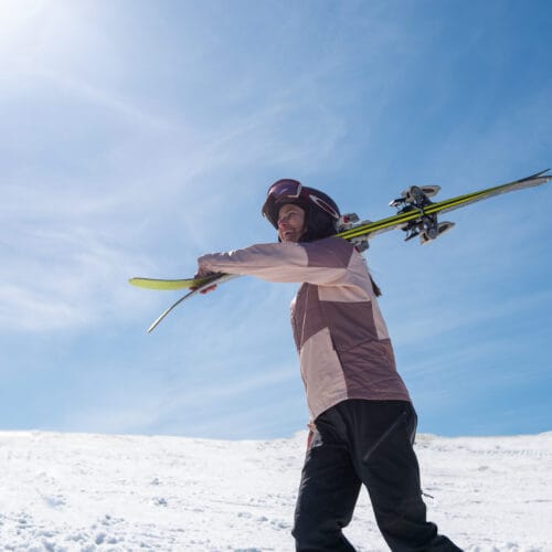 Person bär skidor över axeln på snötäckt fjäll under klarblå himmel.