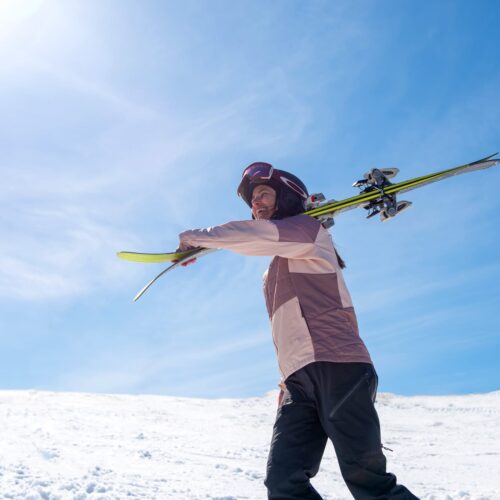 Person carrying skis over shoulder on snowy mountain under clear blue sky
