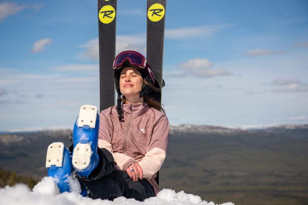 Person sitter i snön med skidor stående bakom och utsikt över fjälllandskap.