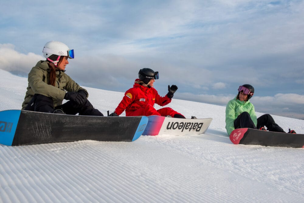 Three snowboarders sitting in snow on a slope with snowboards under a cloudy sky.
