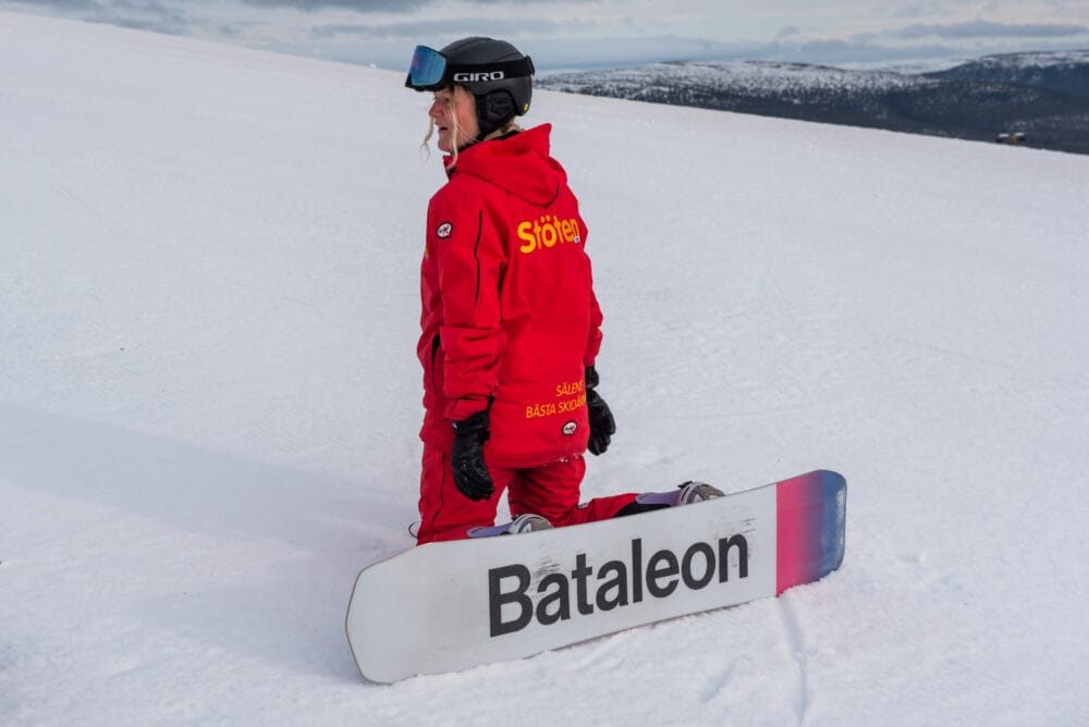 Snowboarder in red jacket kneeling in snow with snowboard and mountain scenery in the background.