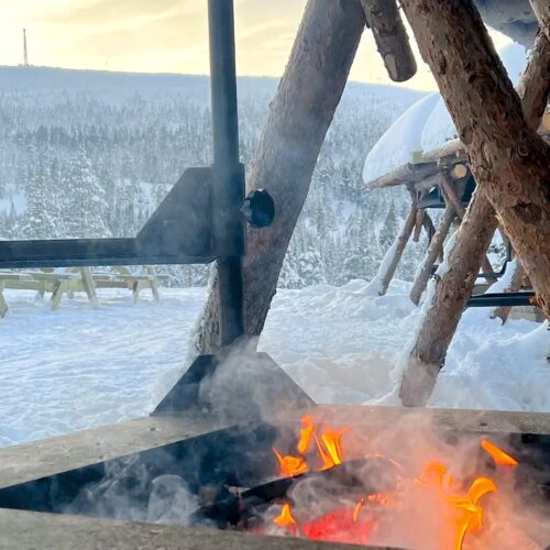 Open fire at shelter with wooden structure, snow-covered tables and mountains in the background
