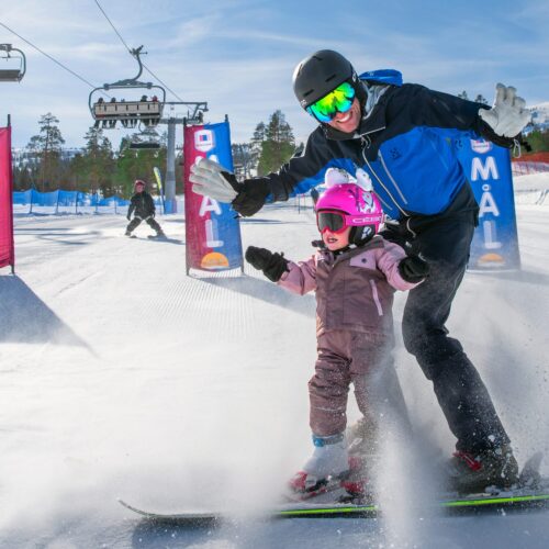 Vuxen hjälper barn åka skidor mellan målflaggor, lekfull fart i solig backe.