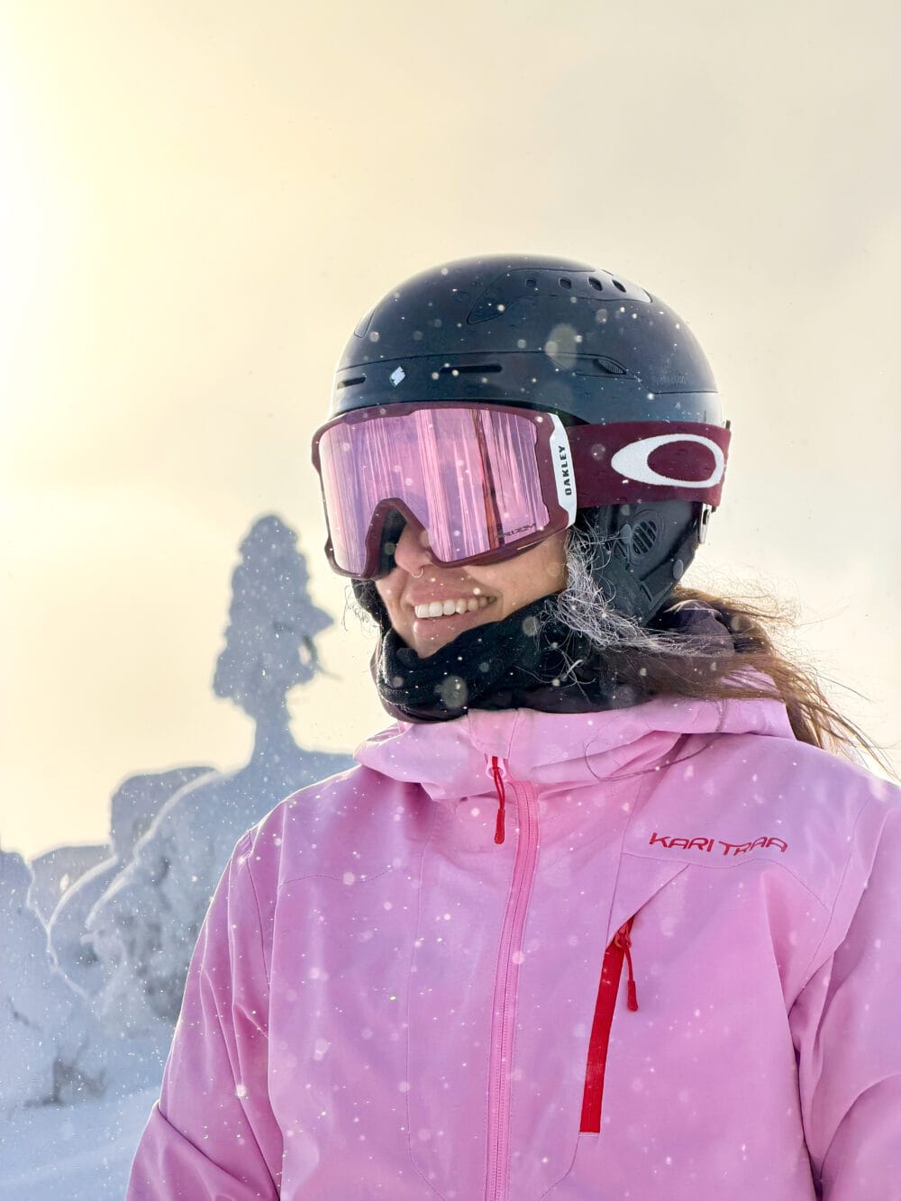 Person i rosa skidjacka med hjälm och goggles står i snöfall mot ljus himmel.