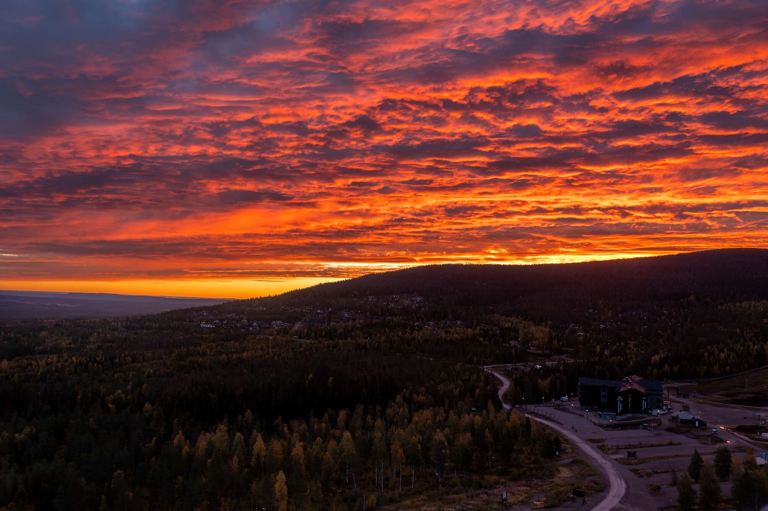 Dramatisk solnedgång med glödande orange moln över skogsklädda kullar och slingrande väg.