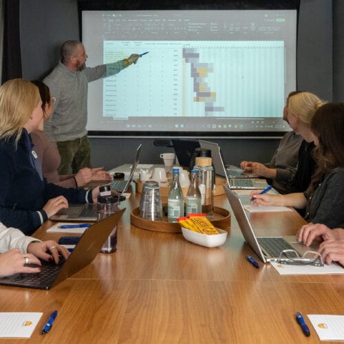 Person peger på projektorskærm med bord og diagrammer, mens flere personer sidder rundt om mødebordet med bærbare computere og noter.