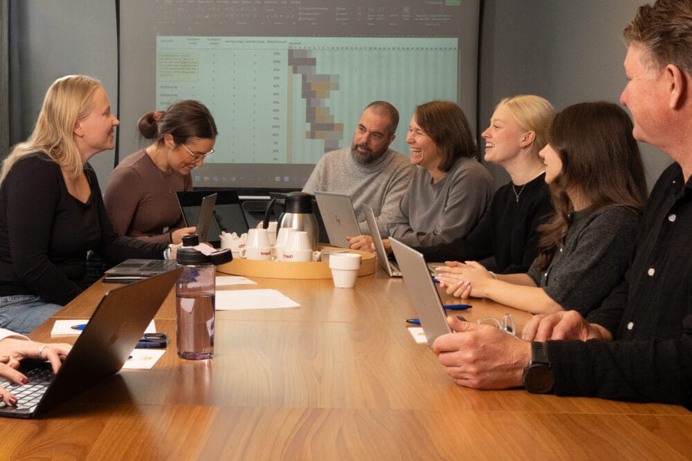 Möte runt stort träbord med laptops, anteckningar och kaffekanna, medan projektorduk visar tabell och färgade diagram