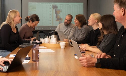 Møde omkring stort træbord med bærbare computere, noter og kaffekande, mens projektorskærmen viser bordet og farvede diagrammer.