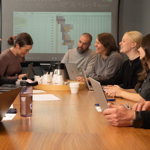 Meeting around large wooden table with laptops, notes and coffee pot, while projector screen shows table and coloured charts