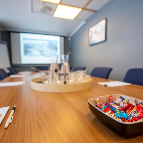 Empty meeting room with long wooden table, notepads and pens, water bottles and coffee cups, as well as a bowl of sweets and a projector screen