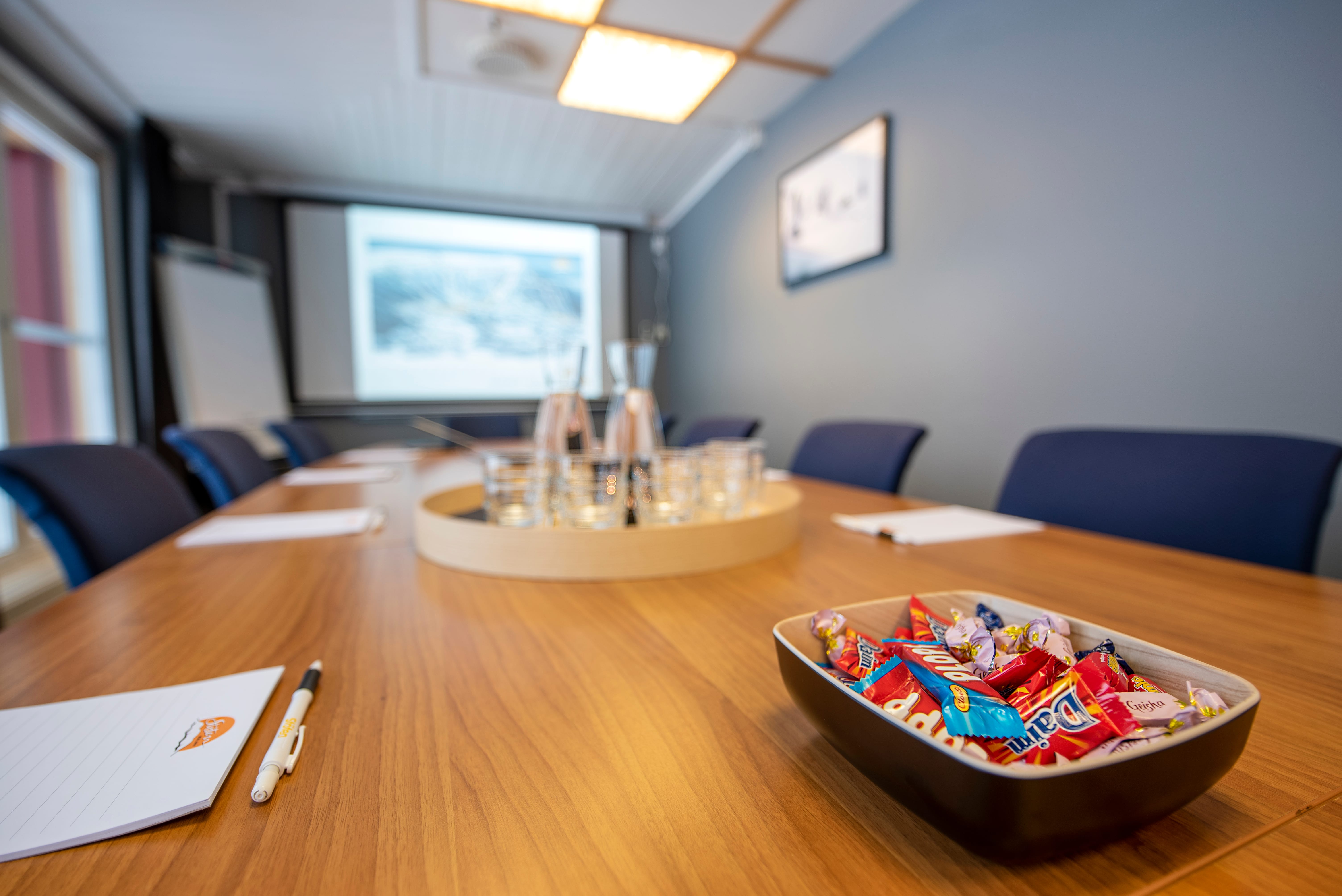 Tomt mötesrum med långt träbord, anteckningsblock och pennor, vattenflaskor och kaffekoppar, samt skål med godis och projektorduk
