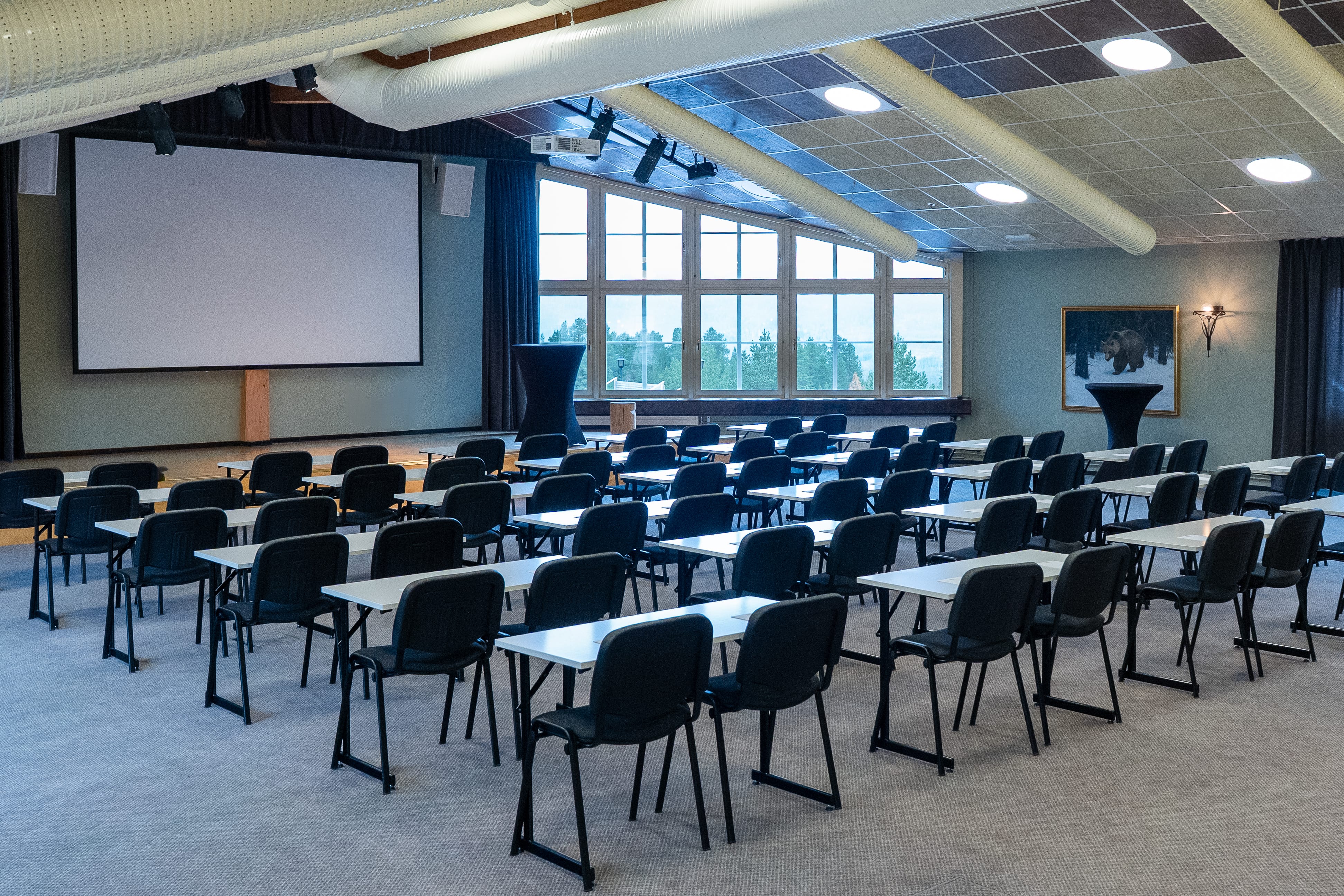 Stort konferensrum med rader av vita bord och svarta stolar, projektorduk längst fram och stora fönster med utsikt mot skog