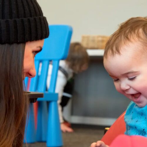 Person med sort kasket læner sig mod børn, der sidder i en rød legetøjsbil indendørs