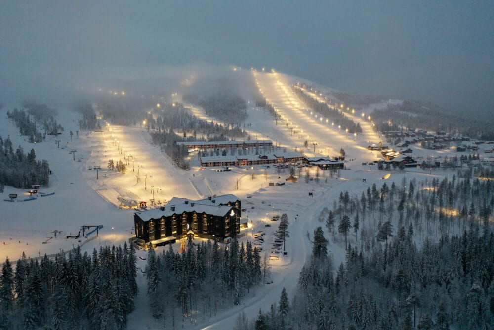 Oplyst skisportssted i skumringen med snedækkede pister, hoteller, veje og omgivende skov i tåge.