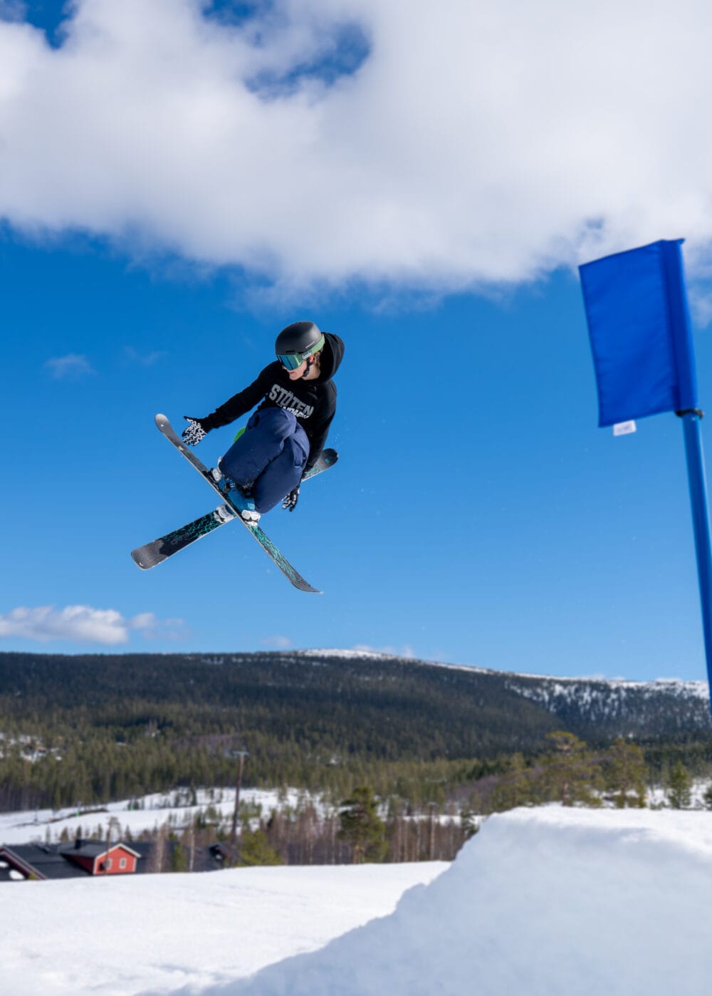 Skidåkare gör hopp i luften över parkhopp med blå himmel och snötäckt fjällbakgrund.