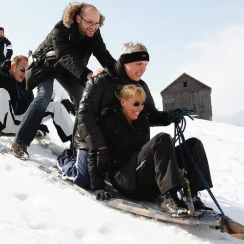 Four people sitting on a large wooden sled on a snowy slope, ready to go down, with a wooden cabin in the background