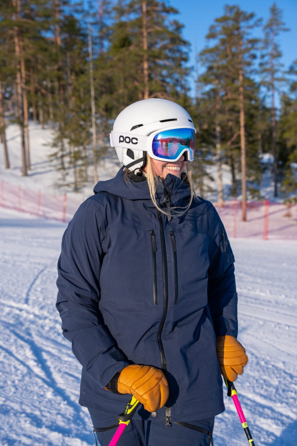Person på skibakke med hvid hjelm, briller og stave, sne og skov i baggrunden.