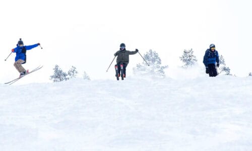 Tre personer åker nedför snötäckt backe, dom hoppar med skidor och svänger i pudersnö