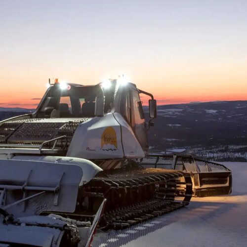 Snow groomer on a groomed ski slope at sunset with mountains and snow-covered landscapes in the background.